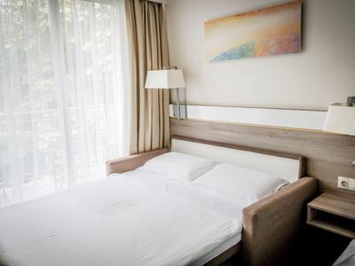 Jasny pokój hotelowy z łóżkiem podwójnym, szafką, lampą stojącą i obrazem na ścianie.