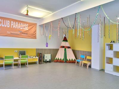 Duży pokój zabaw dla dzieci z kolorowym tipi, ławkami i regałami pod banerem KIDS CLUB PARADISE.