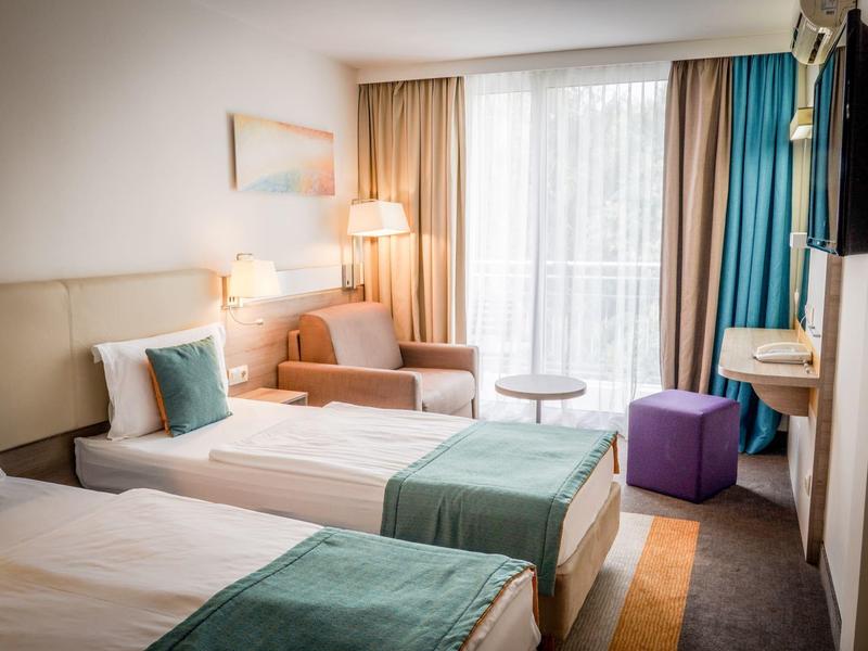 Jasny pokój hotelowy z dwoma łóżkami pojedynczymi, częścią wypoczynkową i balkonem.