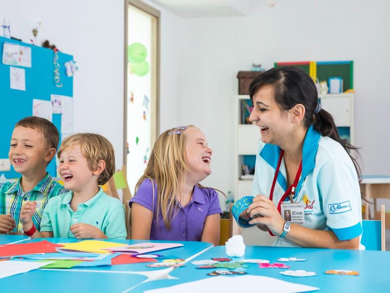 Dzieci śmieją się i tworzą przy stole z opiekunką w jasnym pomieszczeniu.