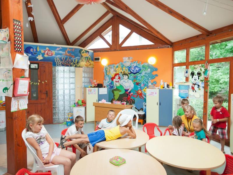 Kolorowe wnętrze przedszkola z kilkorgiem dzieci siedzących przy stołach i krzesłach.