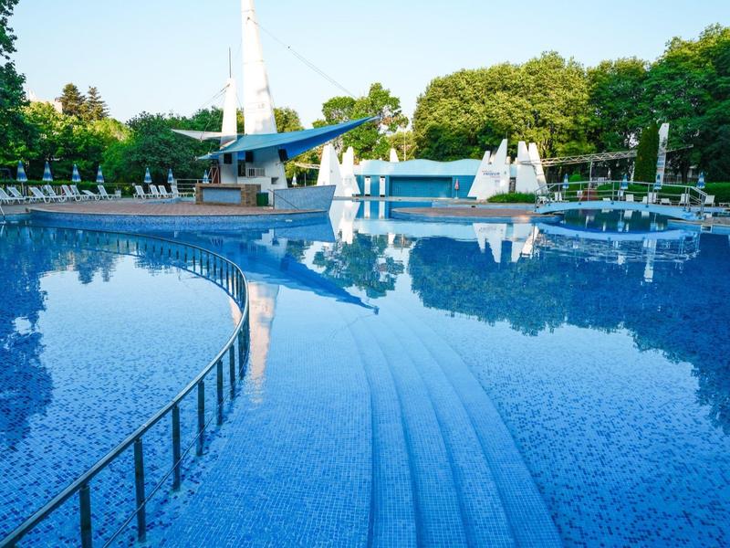 Jasnoniebieski basen hotelowy z schodami i parasolami w zielonym ogrodzie.