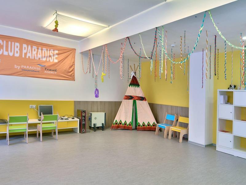 Duży pokój zabaw dla dzieci z kolorowym tipi, ławkami i regałami pod banerem KIDS CLUB PARADISE.
