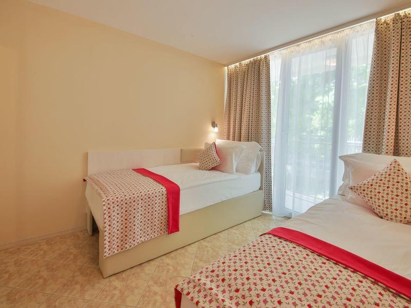 Jasny pokój hotelowy z dwoma łóżkami, czerwonymi akcentami i dużym oknem z zasłonami.