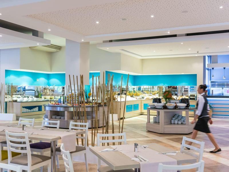 Jasna sala restauracyjna hotelu z białymi stołami i krzesłami, bufetem i obsługą.