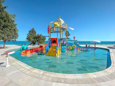 Kolorowy plac zabaw wodny w płytkim basenie z widokiem na plażę i niebieskie niebo.