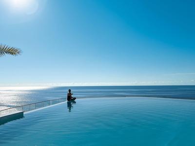 Osoba siedzi przy basenie typu infinity z widokiem na morze pod słonecznym niebem.