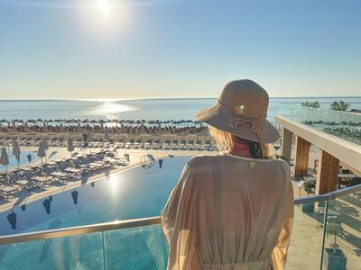 Person stands on balcony overlooking pool and beach at sunset.