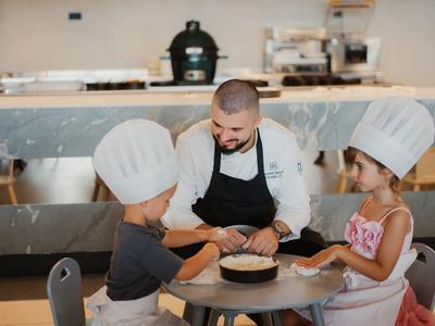 Szef kuchni bawi się z dwójką dzieci w czapkach kucharskich przy małym stoliku w kuchni.