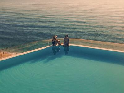 Dwie osoby przy krawędzi basenu infinity z widokiem na spokojne morze o zachodzie słońca.