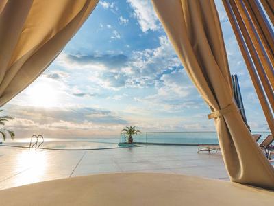 Vista a través de cortinas de una piscina con palmeras y el mar de fondo al atardecer.
