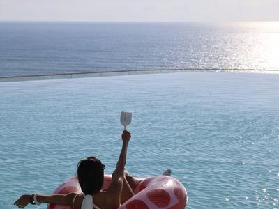 Persona en una piscina sobre un flotador sosteniendo una copa de vino, océano al fondo.