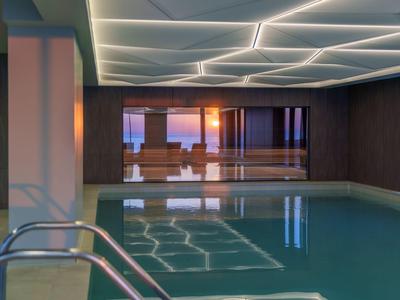 Piscina interior moderna con reflejos y vista al atardecer a través de grandes ventanales.