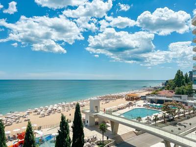 Hotel na plaży z basenem, leżakami i widokiem na morze w słoneczny dzień.