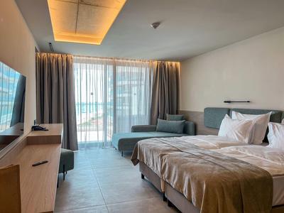 Jasny pokój hotelowy z dużym łóżkiem, sofą, telewizorem i oknem sięgającym podłogi z zasłonami.