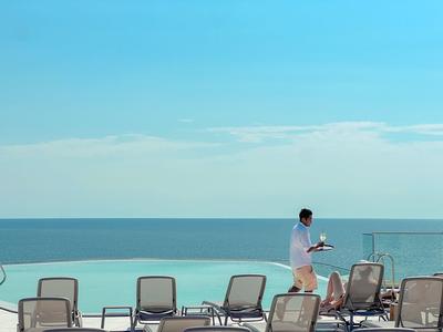 Mężczyzna z tacą obok leżaków przy basenie z widokiem na morze.