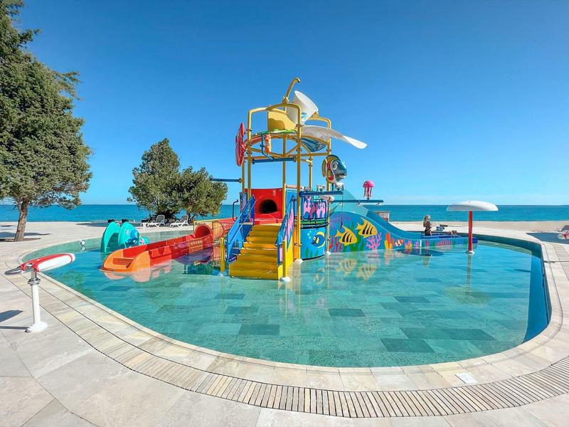 Kolorowy plac zabaw wodny w płytkim basenie z widokiem na plażę i niebieskie niebo.