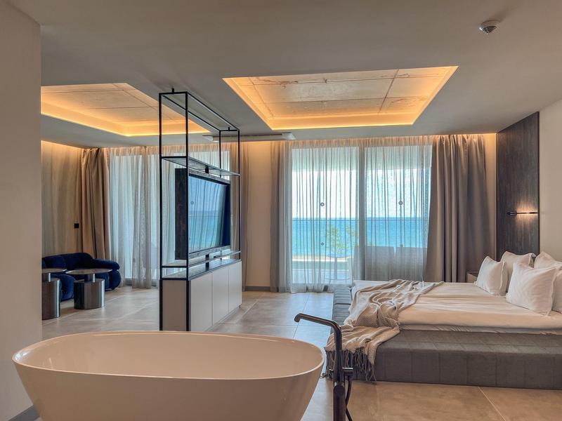 Nowoczesny pokój hotelowy z wolnostojącą wanną, łóżkiem i widokiem na morze przez duże okna.