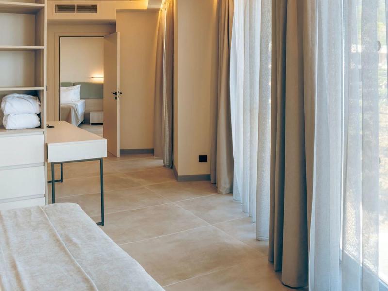 Jasny pokój hotelowy z zasłonami, łóżkiem, wbudowanymi półkami i korytarzem.