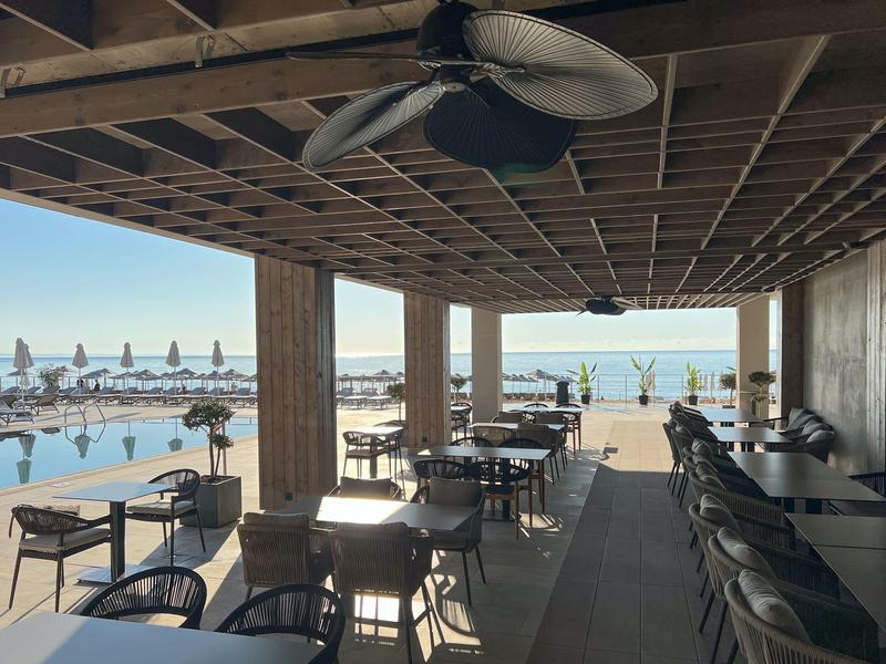 Zadaszony zewnętrzny obszar baru z wentylatorem sufitowym i widokiem na basen i morze.
