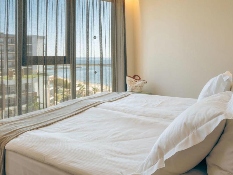 Jasny pokój hotelowy z dużym łóżkiem i widokiem na morze przez okna z zasłonami.