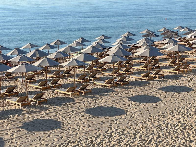Plaża z rzędami drewnianych leżaków pod szarymi parasolami i spokojną wodą w tle