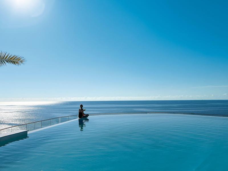 Osoba siedzi przy basenie typu infinity z widokiem na morze pod słonecznym niebem.