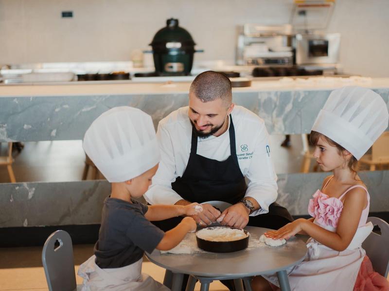 Szef kuchni bawi się z dwójką dzieci w czapkach kucharskich przy małym stoliku w kuchni.