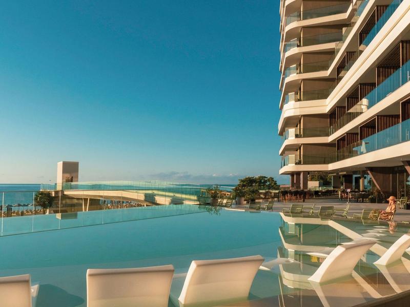Nowoczesny basen infinity z leżakami obok wysokiego budynku hotelowego i widokiem na morze.