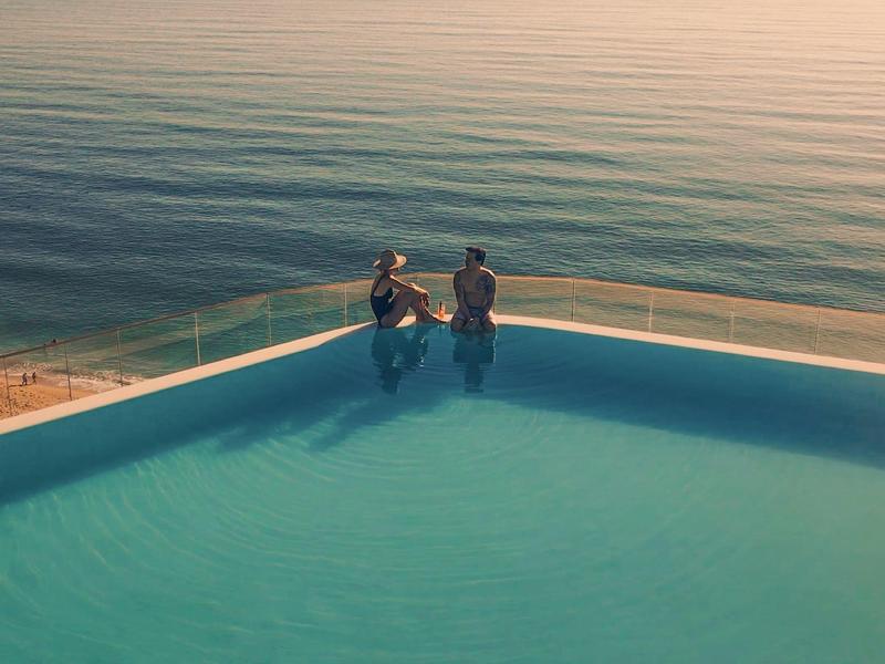 Dwie osoby przy krawędzi basenu infinity z widokiem na spokojne morze o zachodzie słońca.