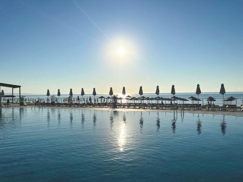 Widok na basen z parasolami i ludźmi na plaży pod słonecznym niebem.