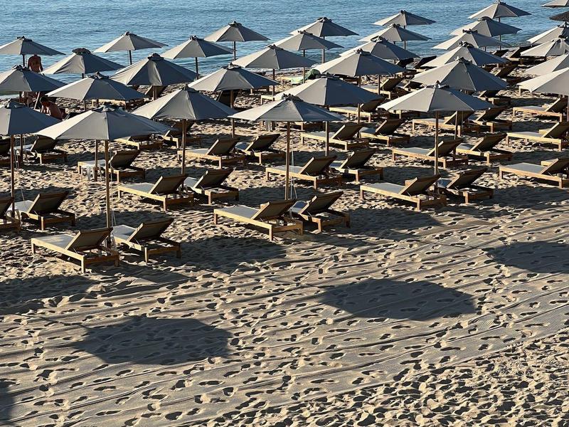 Rząd parasoli i leżaków na piaszczystej plaży z spokojną wodą w tle.