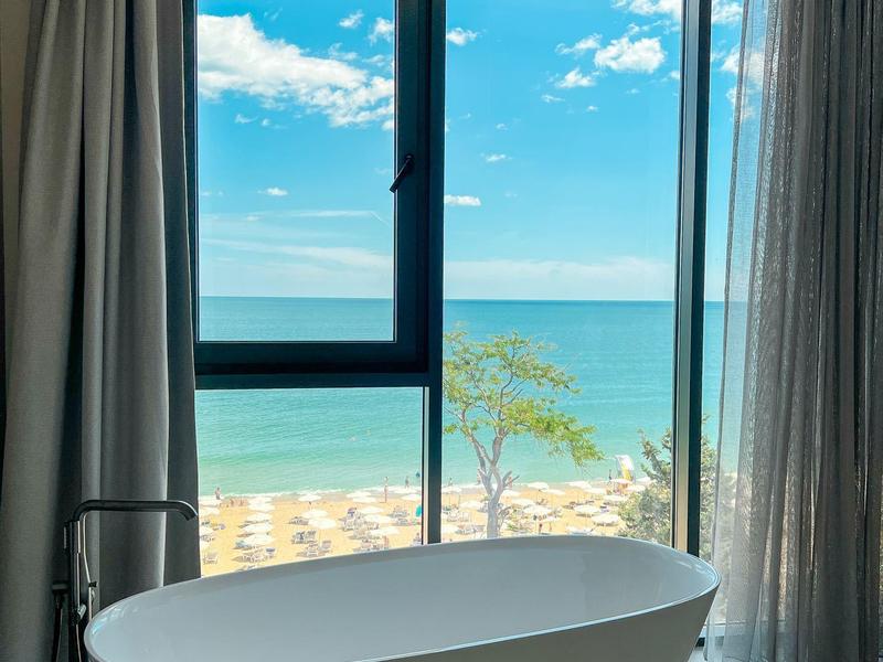 Wolnostojąca wanna przy dużym oknie z widokiem na morze i zasłonami.