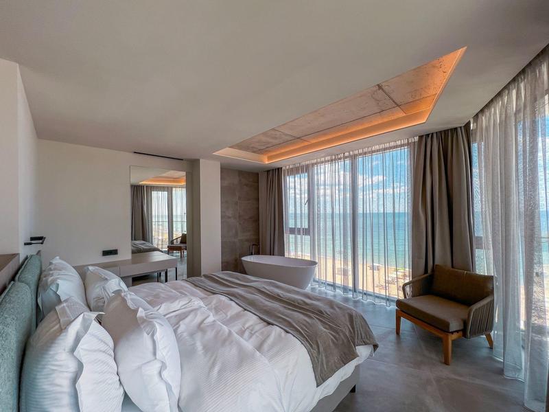 Nowoczesny pokój hotelowy z dużym łóżkiem, wolnostojącą wanną i panoramicznymi oknami z widokiem na morze.