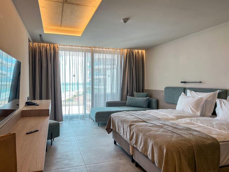 Jasny pokój hotelowy z dużym łóżkiem, sofą, telewizorem i oknem sięgającym podłogi z zasłonami.