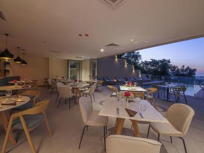 Moderner Innenraum eines Restaurants mit weißen Tischen, Holzmöbeln und Blick auf Pool und Sonnenuntergang.