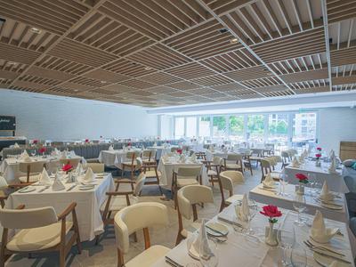 Světlá restaurace s bílými židlemi a prostřenými stoly, velké okno a moderní dřevěný strop.