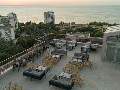 Dachterrasse mit Sitzgruppen, Glasgeländer, Blick auf Bäume, Gebäude und das Meer im Hintergrund.