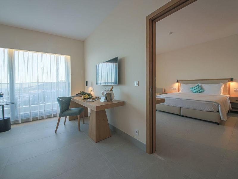 Helles Hotelzimmer mit Balkon, Schreibtisch, Fernseher und angrenzendem Schlafzimmer mit Doppelbett.