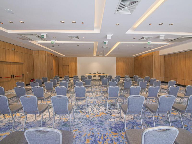 Großer Konferenzraum mit blauen Stühlen, Holzpanelen und hellem, beleuchtetem Deckensystem.