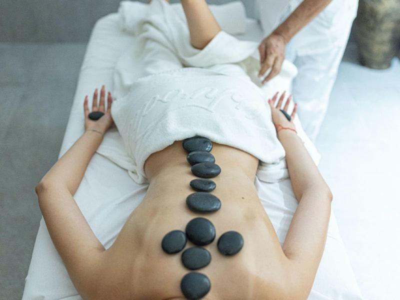 Frau liegt auf Massageliege, Rücken mit heißen Steinen bedeckt, Therapeut hält Arm zur Behandlung.