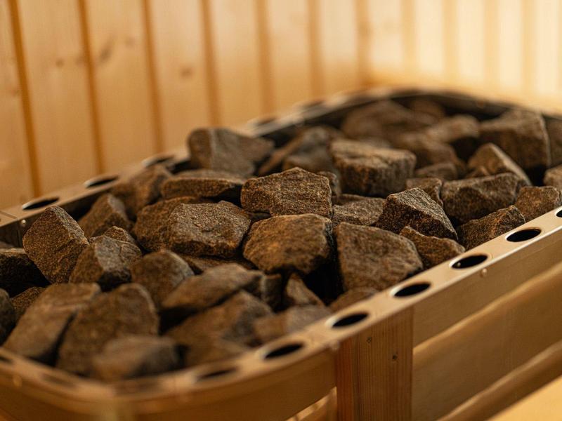 Metallbehälter mit dunklen Saunasteinen in Holzverkleidung, warme Beleuchtung, rustikaler Stil.