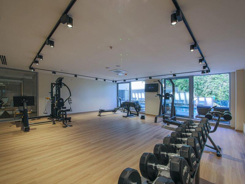 Moderner Fitnessraum mit Holzfußboden, Hanteln, Geräten und großen Fenstern für Tageslicht.