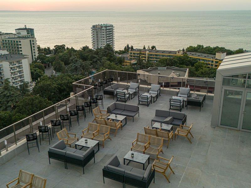 Dachterrasse mit Sitzgruppen, Glasgeländer, Blick auf Bäume, Gebäude und das Meer im Hintergrund.