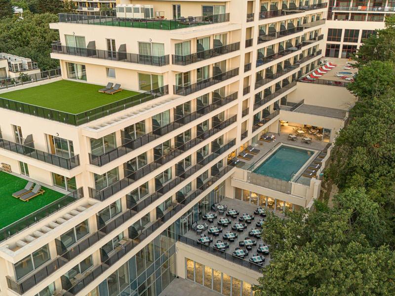 Modernes, weißes Wohnhaus mit großen Balkonen, grünen Dachterrassen und kleinem Pool neben Waldgebiet.