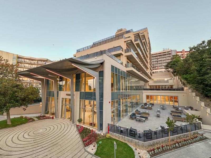 Moderne mehrstöckige Glas- und Betongebäude mit Terrasse, Garten und Außenbestuhlung bei klarem Himmel.