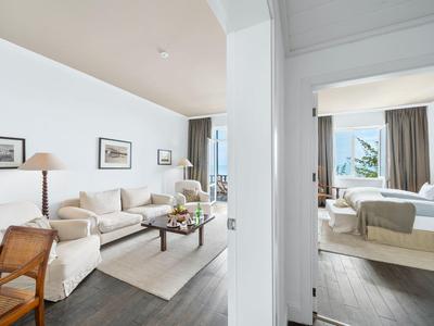 Camera d'hotel luminosa con zona giorno, divano, quadri alle pareti e camera da letto adiacente con vista mare.