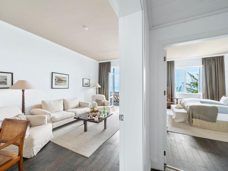 Camera d'hotel luminosa con zona giorno, divano, quadri alle pareti e camera da letto adiacente con vista mare.