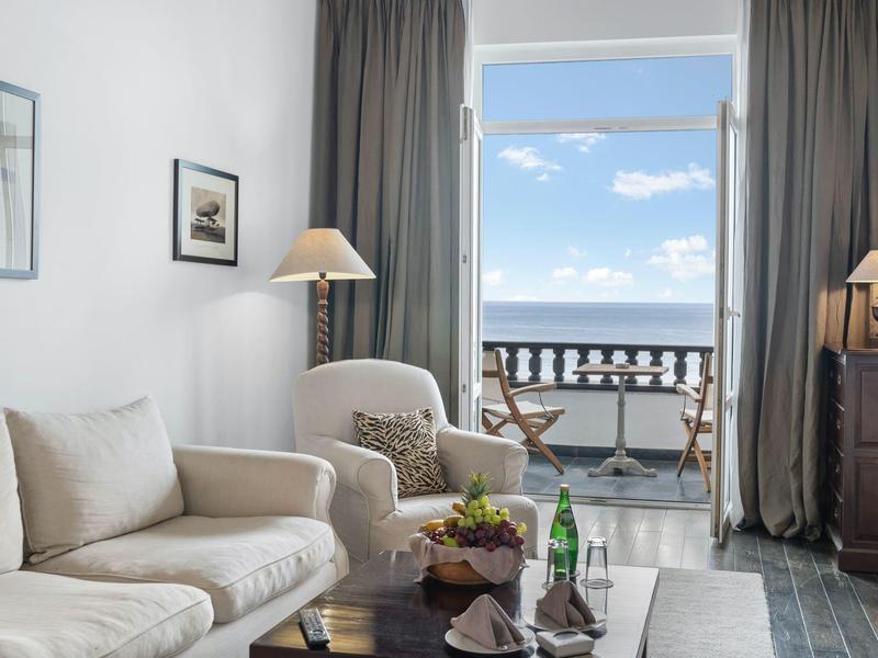 Soggiorno luminoso con divano beige, poltrona, lampade e balcone con vista sul cielo azzurro.