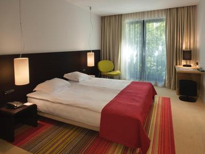 Nowoczesny pokój hotelowy z dwoma pojedynczymi łóżkami, czerwonym narzutem, fotelem i biurkiem przy oknie.